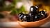 Un cucchiaio di legno contenente diverse olive nere immerse nell'olio d'oliva, con uno sfondo sfocato. - Olive Oil Times