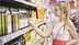 Una mujer de pelo largo con un vestido blanco y rosa busca una botella de aceite de oliva en el estante de un supermercado. Olive Oil Times