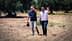 Deux hommes marchant dans une oliveraie, l'un faisant signe de la main et l'autre se protégeant les yeux du soleil. Olive Oil Times