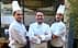 Three chefs wearing white uniforms and hats standing in front of a restaurant entrance. - Olive Oil Times