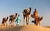 Two men walking alongside camels in a desert environment, with saddles and bags on the camels' backs. - Olive Oil Times