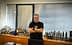 Uomo con i capelli grigi che indossa una maglietta nera in piedi in un ufficio con prodotti a base di olio d'oliva esposti su uno scaffale. - Olive Oil Times