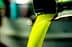 Un chorro de aceite de oliva de color verde brillante se vierte desde un pico metálico hacia un recipiente. Olive Oil Times