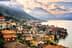 Lakeside village with houses and mountains in the background under a cloudy sky. - Olive Oil Times
