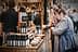 Various bottles of olive oil displayed on a wooden table at a market with people in the background. - Olive Oil Times