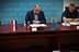 A person sitting at a table with flags of Croatia and another entity displayed on stands. - Olive Oil Times