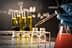 A laboratory scene showing a hand in a blue glove using a pipette to transfer liquid into small glass bottles. - Olive Oil Times