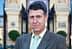 A man wearing a brown suit and a light green tie, standing outdoors in front of a building. - Olive Oil Times