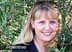A woman with blonde hair smiling while standing among green leaves and branches. - Olive Oil Times