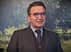Homme portant un costume et des lunettes, debout devant un fond texturé avec de la verdure. - Olive Oil Times