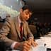 A man in a suit signing documents during a conference with an audience in the background. - Olive Oil Times