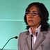 Woman with glasses speaking at a podium during a presentation with a green background. - Olive Oil Times