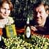 Two individuals posing with bottles of olive oil and a crate of olives in a field. - Olive Oil Times