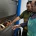 Two men inspecting a baking oven with bread inside, one wearing an apron and the other in a green shirt. - Olive Oil Times
