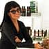 Woman with dark hair and sunglasses smiling while leaning against a counter with products in the background. - Olive Oil Times