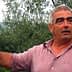 Man with gray hair gesturing while standing in an olive grove with trees in the background. - Olive Oil Times