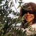 Woman smiling while harvesting olives from an olive tree in a sunny outdoor setting. - Olive Oil Times