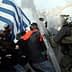 A protest scene in Greece featuring a person holding a Greek flag and police in riot gear. - Olive Oil Times