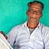 An elderly Indian farmer sitting against a turquoise wall, wearing a white shirt and pants. - Olive Oil Times