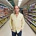 A man in a striped shirt standing in the aisle of a grocery store surrounded by various packaged products. - Olive Oil Times