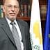 A man in a gray suit standing next to a flag of Cyprus and the European Union flag. - Olive Oil Times
