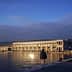 Trieste Maritime Station building reflecting on the water during sunset with a person walking nearby. - Olive Oil Times