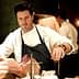 Chef in a white coat and apron preparing a piece of meat in a kitchen setting. - Olive Oil Times