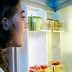 A person with dark hair looking into an open refrigerator filled with various food items and a bottle of oil. - Olive Oil Times