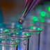 A pipette dropping liquid into a row of clear test tubes illuminated with green light. - Olive Oil Times