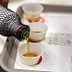 A bottle pouring olive oil into small tasting cups on a tray. - Olive Oil Times