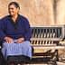 Older woman wearing a blue sweater and skirt sitting on a wooden bench against a wall. - Olive Oil Times