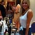 Woman with long blonde hair standing at a table filled with various product bottles at an event. - Olive Oil Times