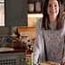 Woman smiling while holding a plate of toast next to a bottle of Carbonell olive oil in a kitchen. - Olive Oil Times