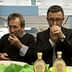 Two men tasting olive oil from small cups at an event, focusing on the aroma and flavor. - Olive Oil Times