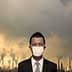 Businessman in a suit and mask standing against an industrial backdrop with smoke stacks and cloudy sky. - Olive Oil Times