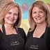 Two women wearing black aprons with 'Cleo's' logo, smiling at the camera in a studio setting. - Olive Oil Times