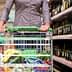Person pushing a shopping cart filled with groceries in a supermarket aisle with shelves of bottles. - Olive Oil Times