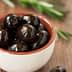 A small bowl filled with black olives placed on a wooden surface. - Olive Oil Times