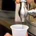 A hand operating a metal oil dispenser to fill a clear plastic cup with dark liquid. - Olive Oil Times