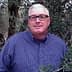 Man wearing glasses and a blue checkered shirt standing among olive trees. - Olive Oil Times
