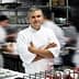 A chef standing with arms crossed in a busy professional kitchen environment. - Olive Oil Times