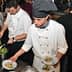 Two chefs in white uniforms plating food in a busy kitchen environment. - Olive Oil Times
