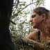 A woman with curly hair sitting among olive tree branches, looking thoughtfully to the side. - Olive Oil Times