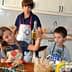 Three children in a kitchen, one holding a bottle of olive oil while others observe. - Olive Oil Times