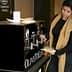 A woman dispensing olive oil from a black dispenser labeled 'OLIVETO LIVE' in a casual setting. - Olive Oil Times