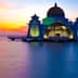 A mosque with a golden dome and colorful lighting situated by the water during sunset. - Olive Oil Times