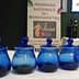Four blue glass jars arranged in a row at an olive oil event with a promotional banner in the background. - Olive Oil Times