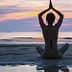 Individual sitting in a yoga pose on the beach during sunset with hands raised. - Olive Oil Times