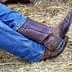 A pair of brown cowboy boots worn by a person seated, with blue jeans visible. - Olive Oil Times