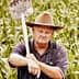 A man wearing a hat and overalls holding a shovel in a cornfield. - Olive Oil Times